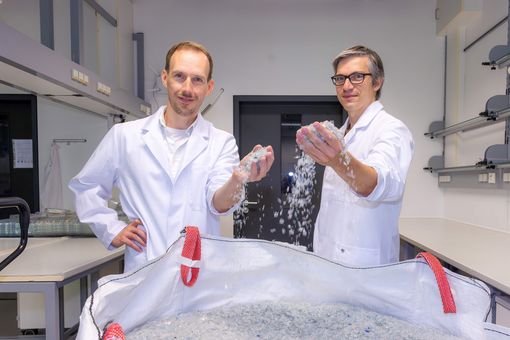 Dr. Ronny Frank und Dr. Christian Sonnendecker stehen vor einem Sack voll kleingehäckselten Plastikteilen. 